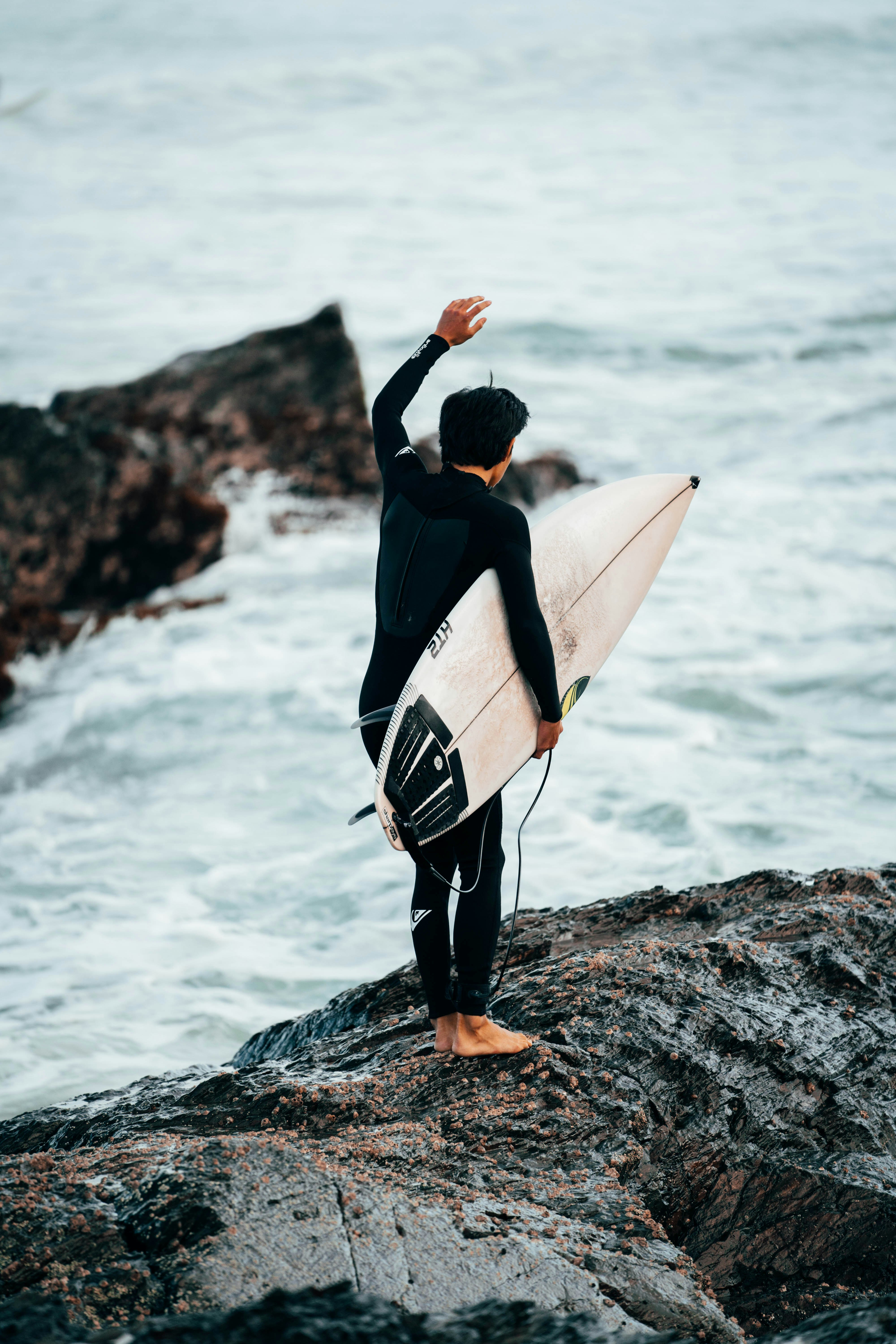 man in black long sleeve shirt carrying white surfboard standing on rocky shore during daytime